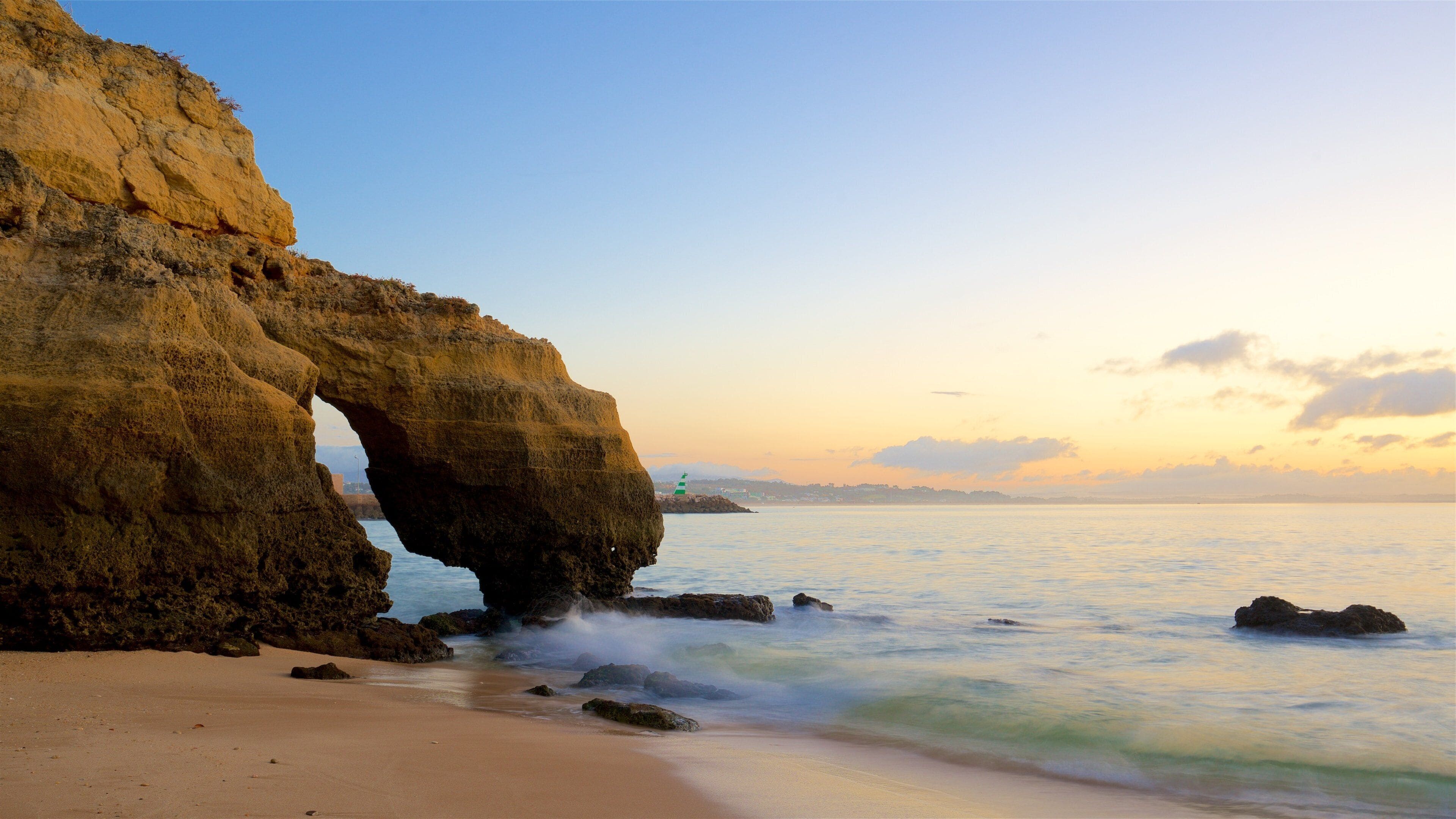 A coastal landscape with a large rock arch formation, sandy beach, and calm sea.