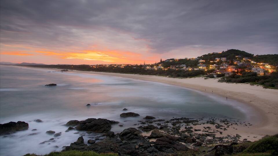A coastal sunset with a beach, rocky shoreline, and a hillside town with houses.