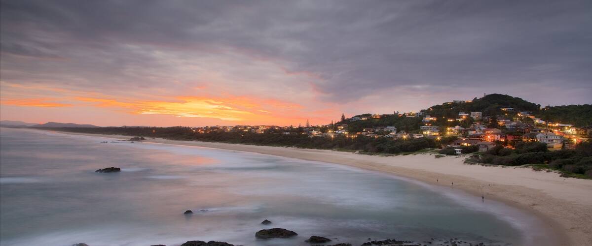 A coastal sunset with a beach, rocky shoreline, and a hillside town with houses.