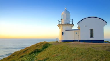 Um farol com estrutura branca e azul, uma colina coberta de grama e o oceano ao fundo.