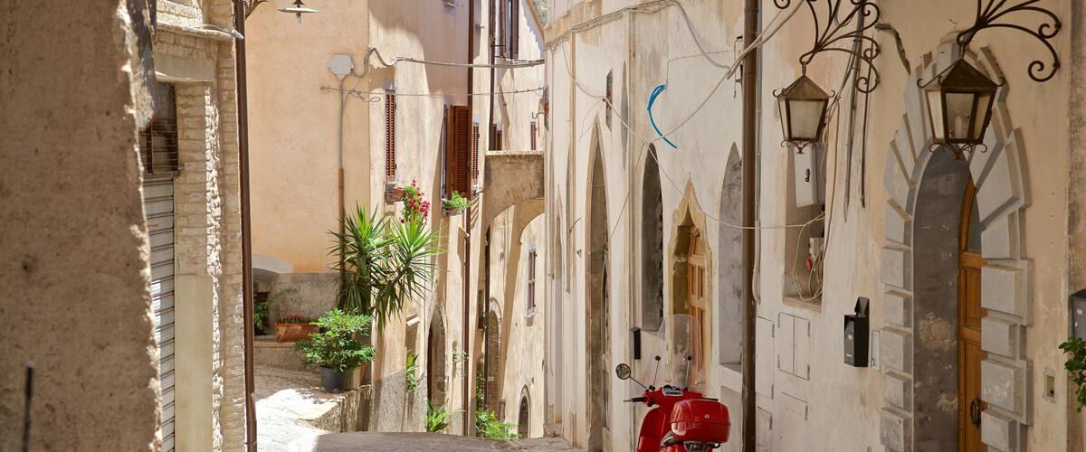 A narrow cobblestone street with a red scooter parked on the right, flanked by buildings with arched doorways and hanging lanterns.