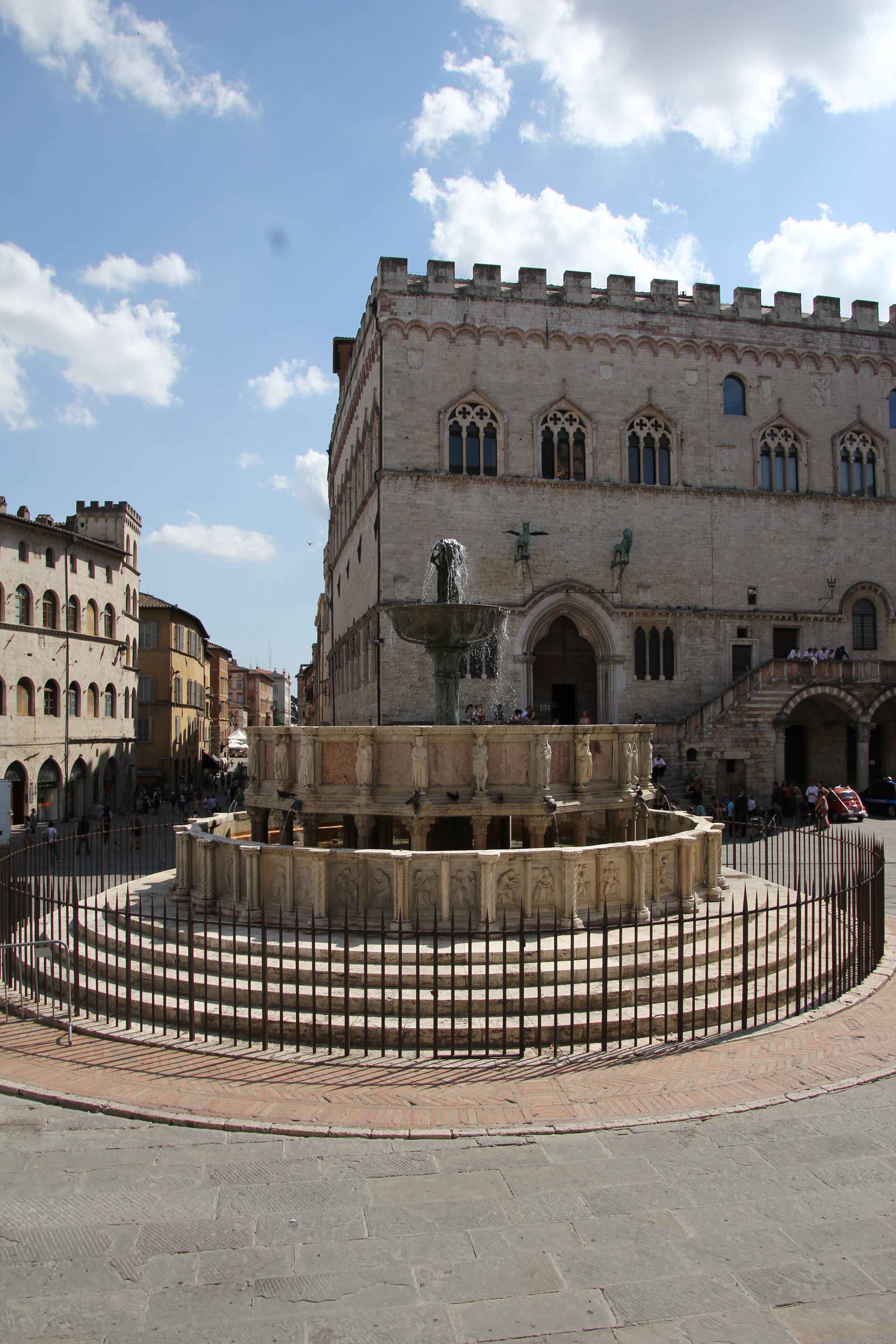 A historic fountain with statues in the center, surrounded by a stone staircase and a metal fence. In the background, there is a large building with arched windows and a clock tower.