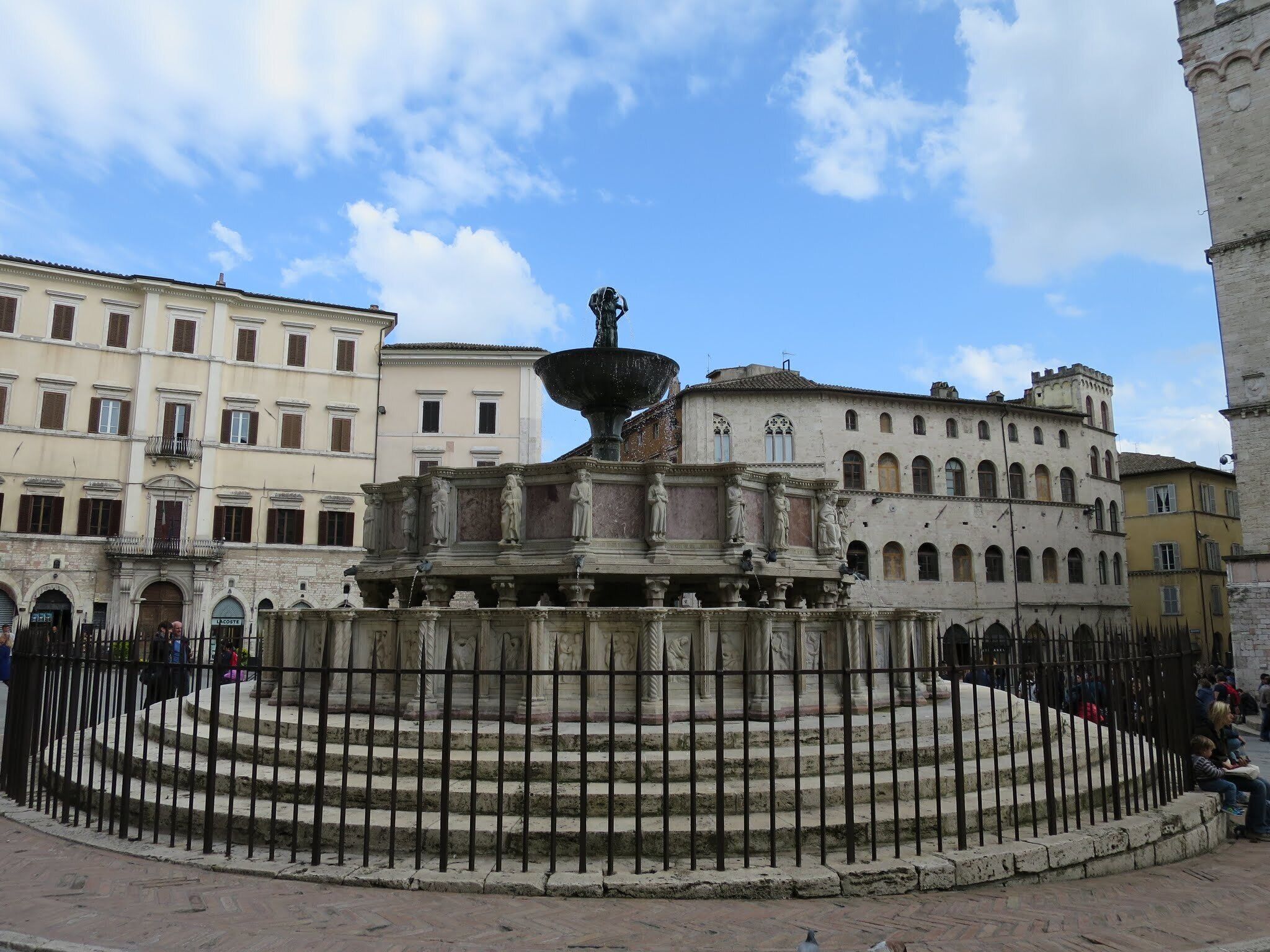 Une fontaine historique, ornée de statues, se trouve sur une place entourée d’édifices à l’architecture classique.