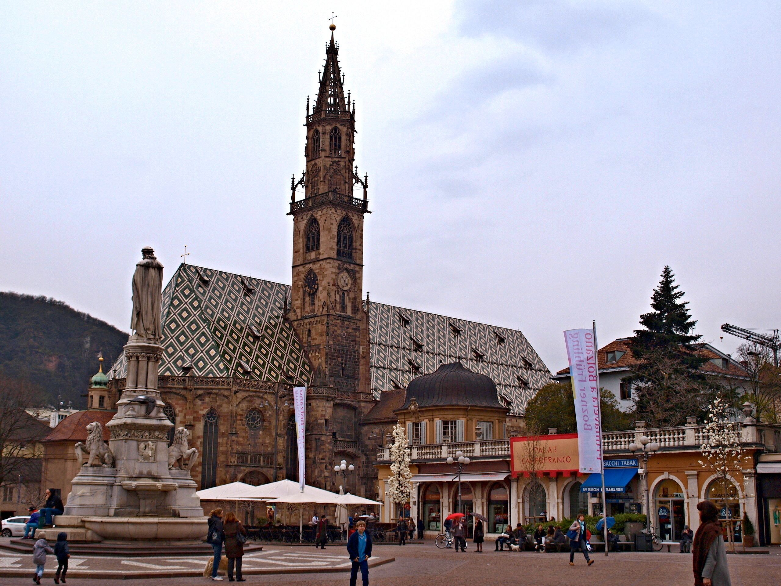 Ein historischer Platz mit einem markanten Kirchturm, einer Statue und einem Brunnen.