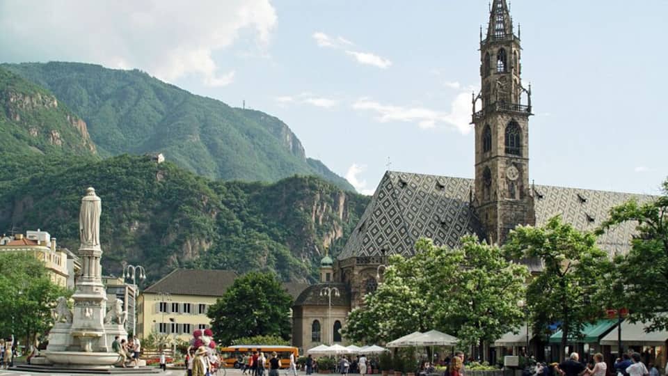 A town square with a prominent clock tower, a statue, and a mountain backdrop.