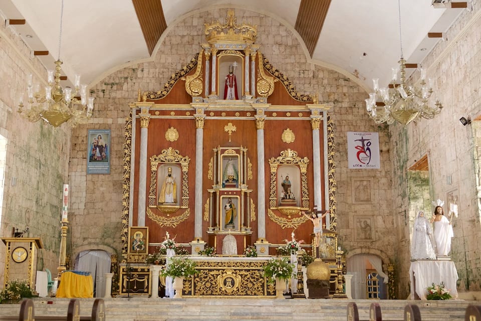 Ein kunstvoll gestaltetes Kircheninnere mit einem zentralen Altar, religiösen Statuen und einer Wand aus Stein im Hintergrund.