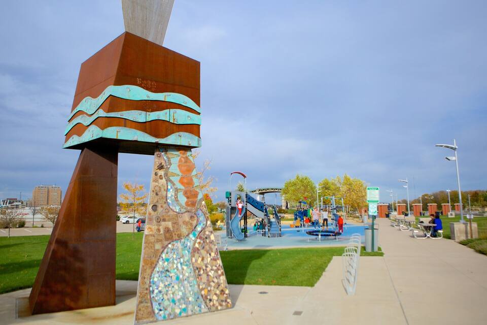 A playground with slides and climbing structures, surrounded by a park with a large sculpture and a clear sky.
