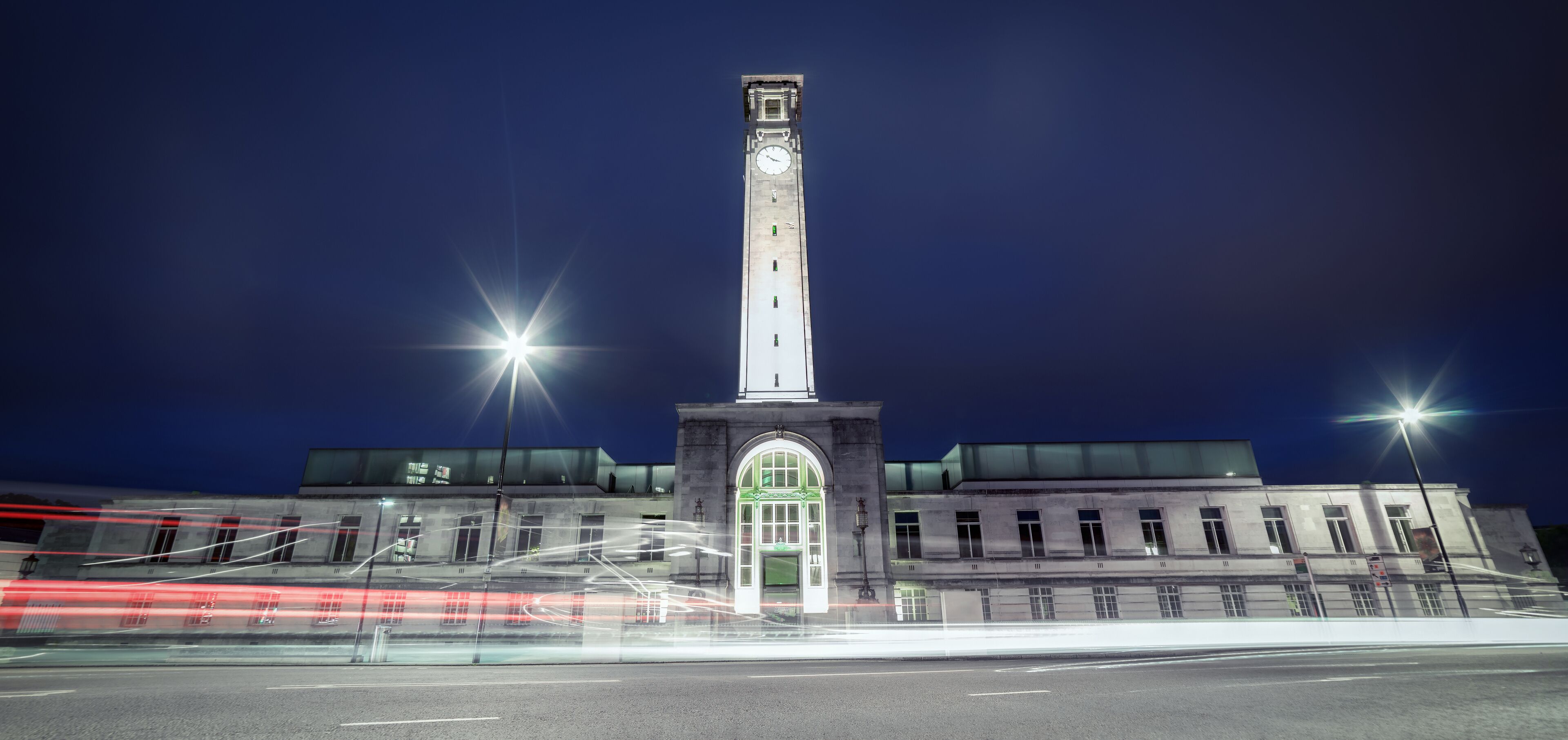 Une haute tour d’horloge illuminée la nuit, avec des lampadaires et des traînées de lumière laissées par les véhicules en mouvement.