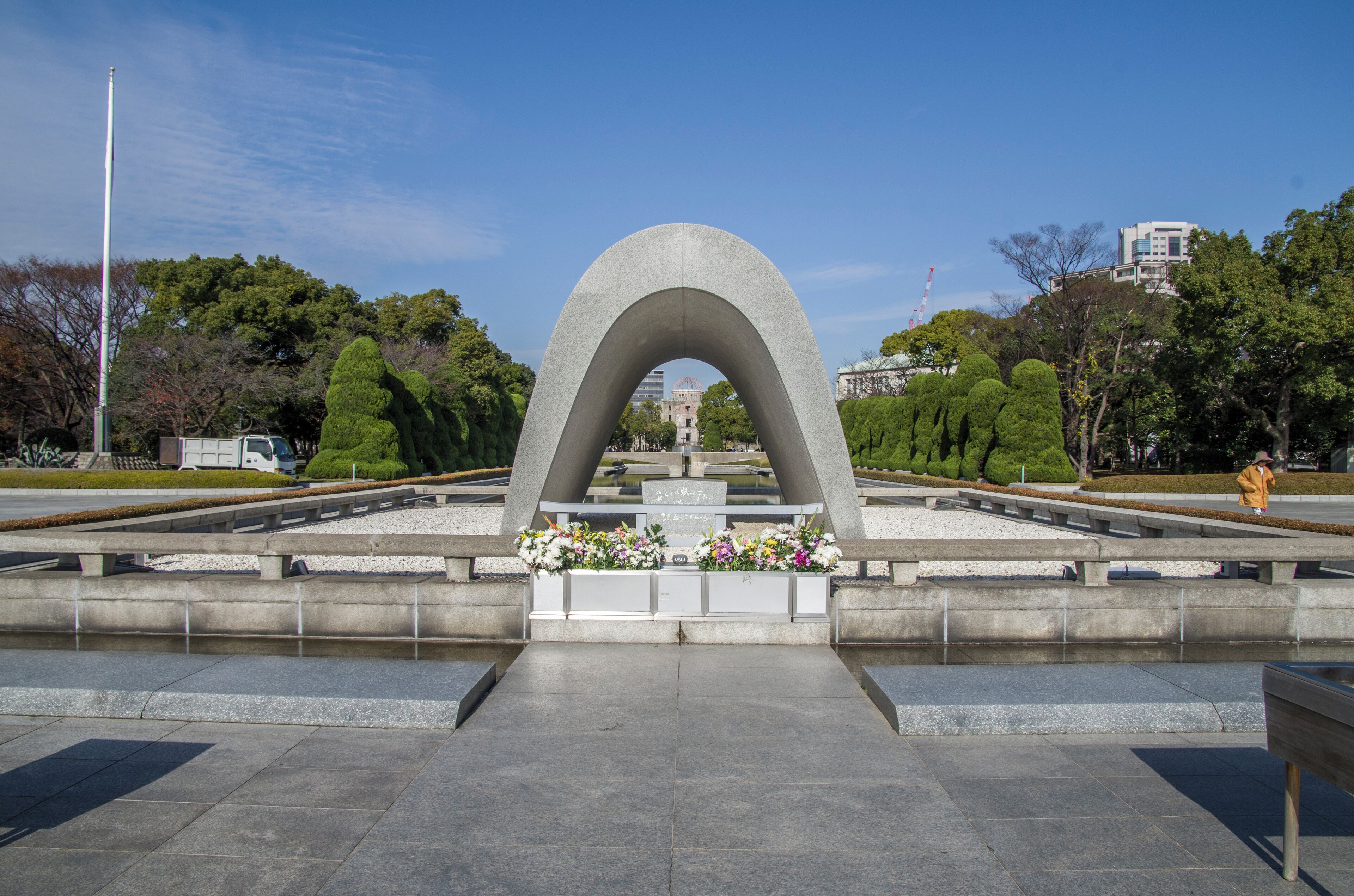 A large concrete arch monument with floral arrangements at its base, surrounded by a park with trees and a distant building.