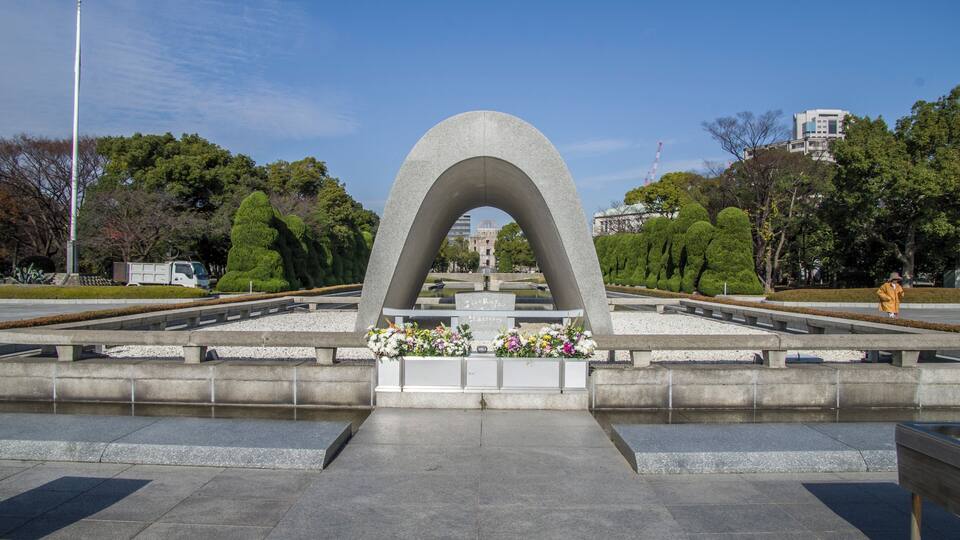 A large concrete arch monument with floral arrangements at its base, surrounded by a park with trees and a distant building.