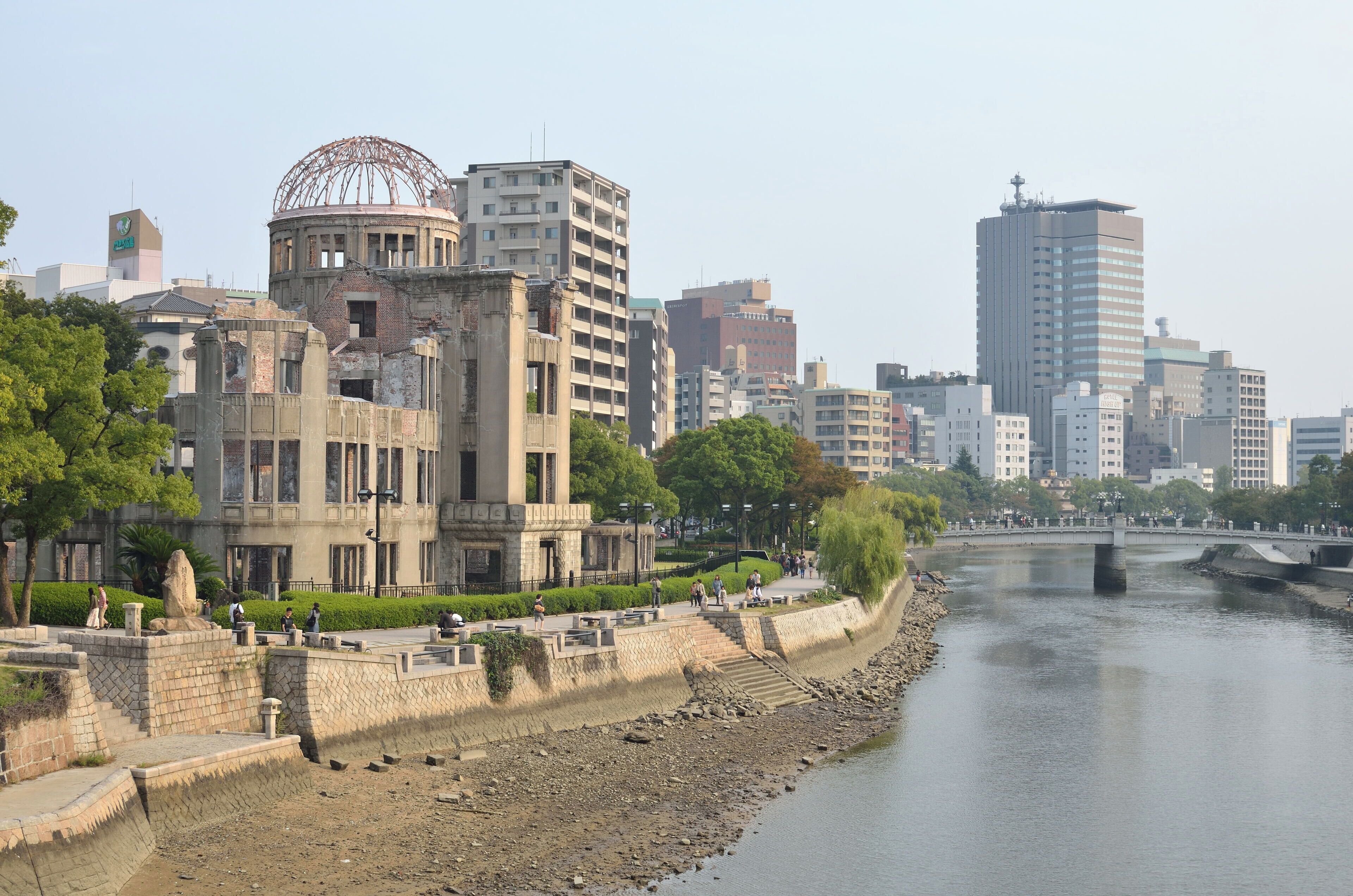 A riverside cityscape with historic and modern buildings, a bridge, and a park with people walking.