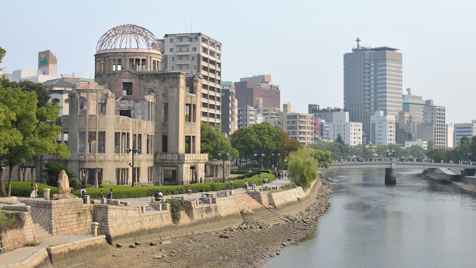 A riverside cityscape with historic and modern buildings, a bridge, and a park with people walking.