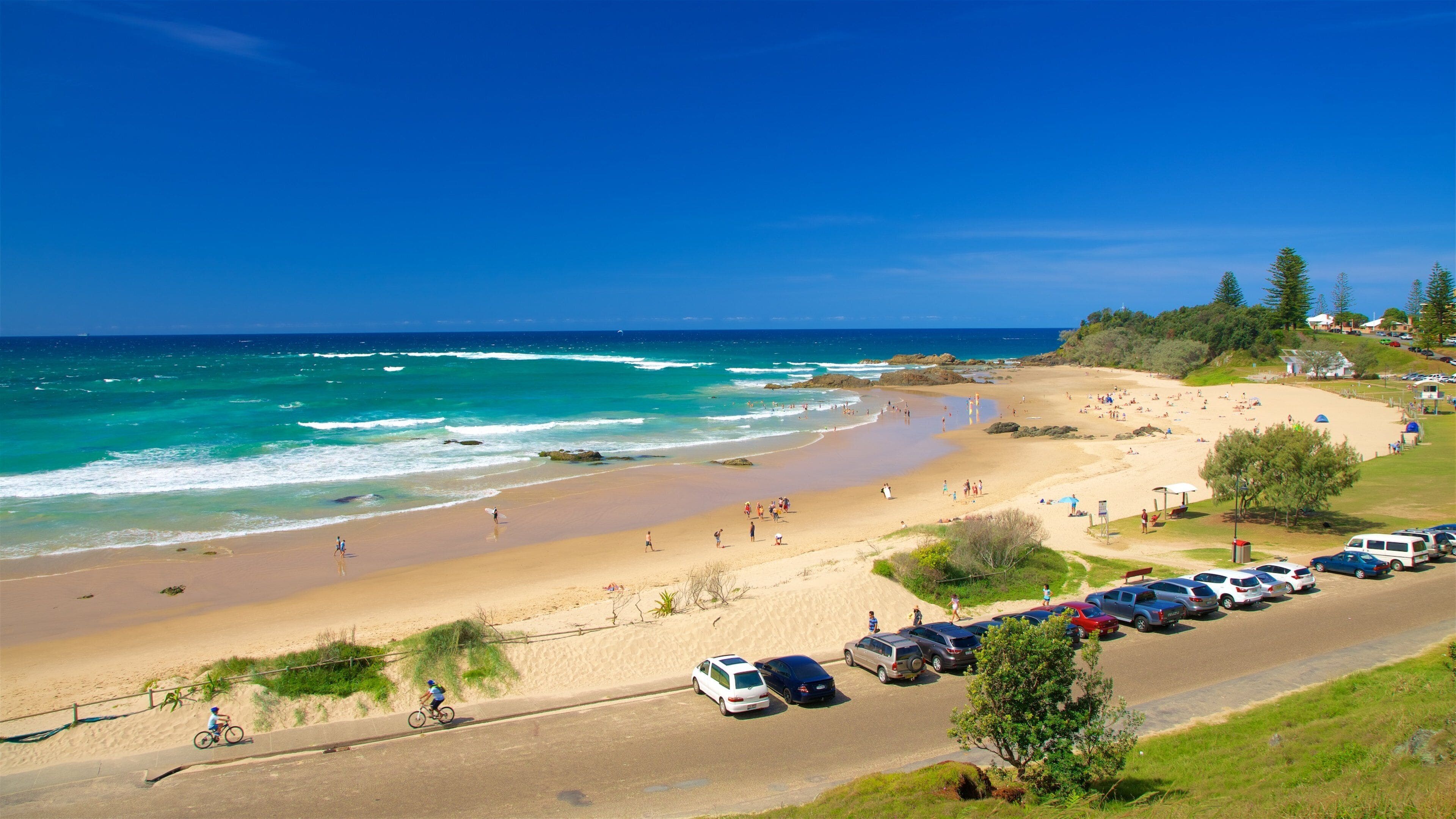 Une plage aux eaux d’un bleu limpide, avec un rivage sablonneux et des voitures garées le long de la route.