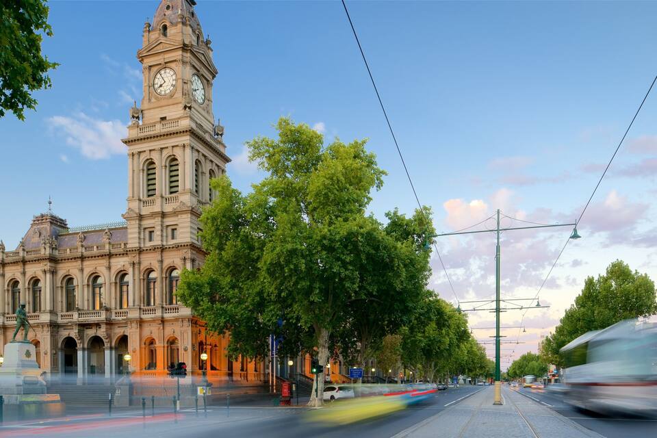 A historic building with a clock tower, surrounded by trees and a tram line.