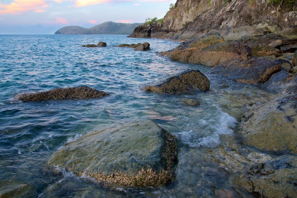 Escena costera con rocas grandes al frente y un acantilado rocoso al fondo.