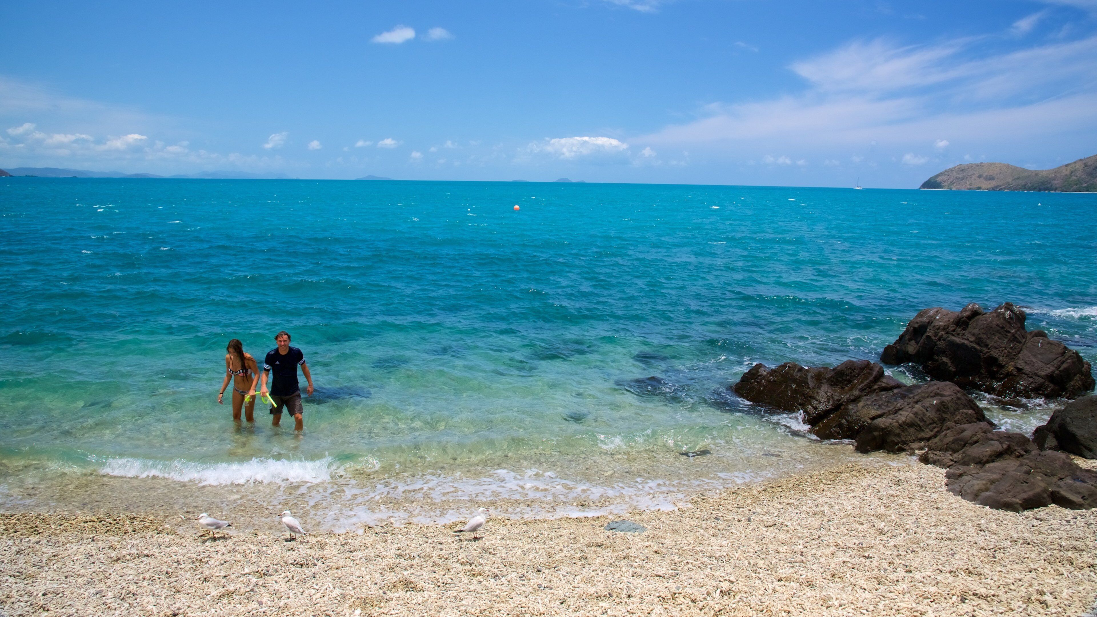 Una playa con aguas turquesas cristalinas, costa rocosa y montañas a lo lejos.