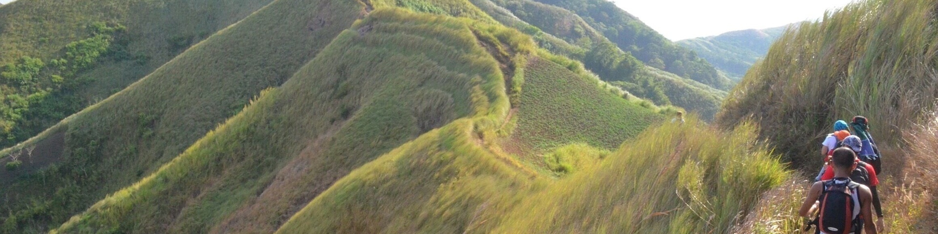 A hiker on a narrow trail surrounded by lush green hills.