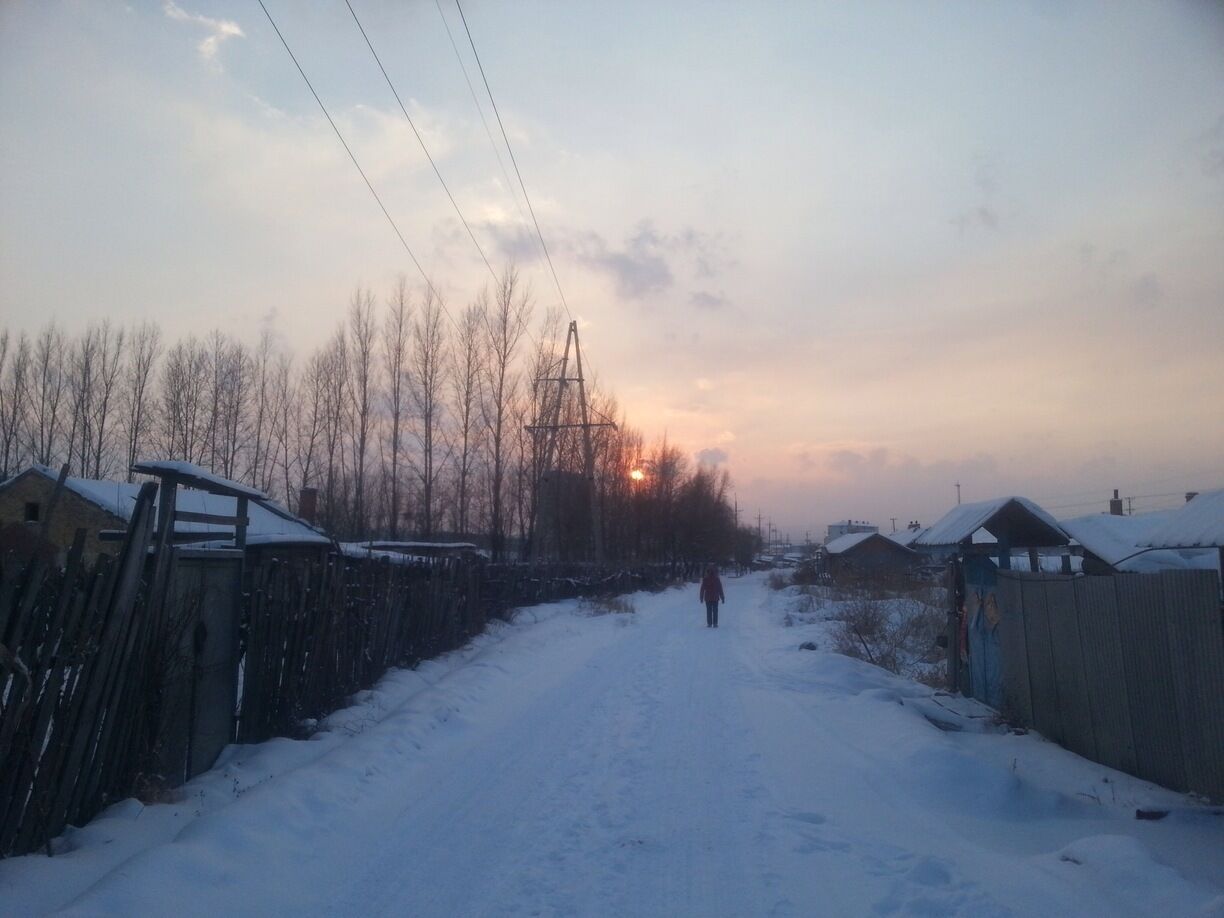 A snow-covered rural road flanked by bare trees and wooden fences under a sunset sky.