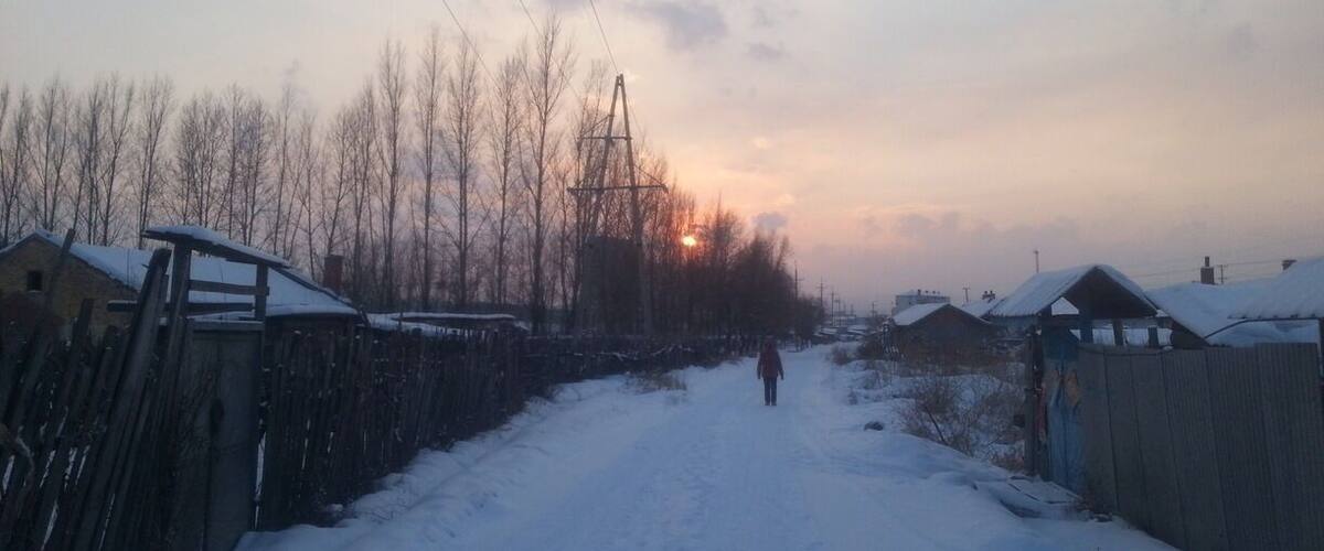 A snow-covered rural road flanked by bare trees and wooden fences under a sunset sky.