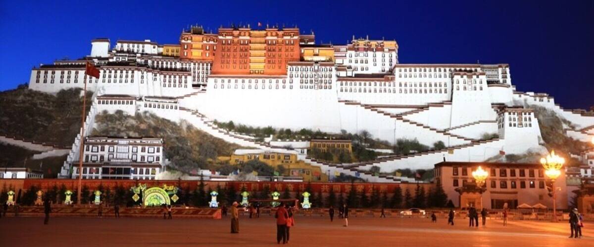 A large, illuminated fortress-like structure with multiple levels and a prominent red building on top, surrounded by a hilly landscape.