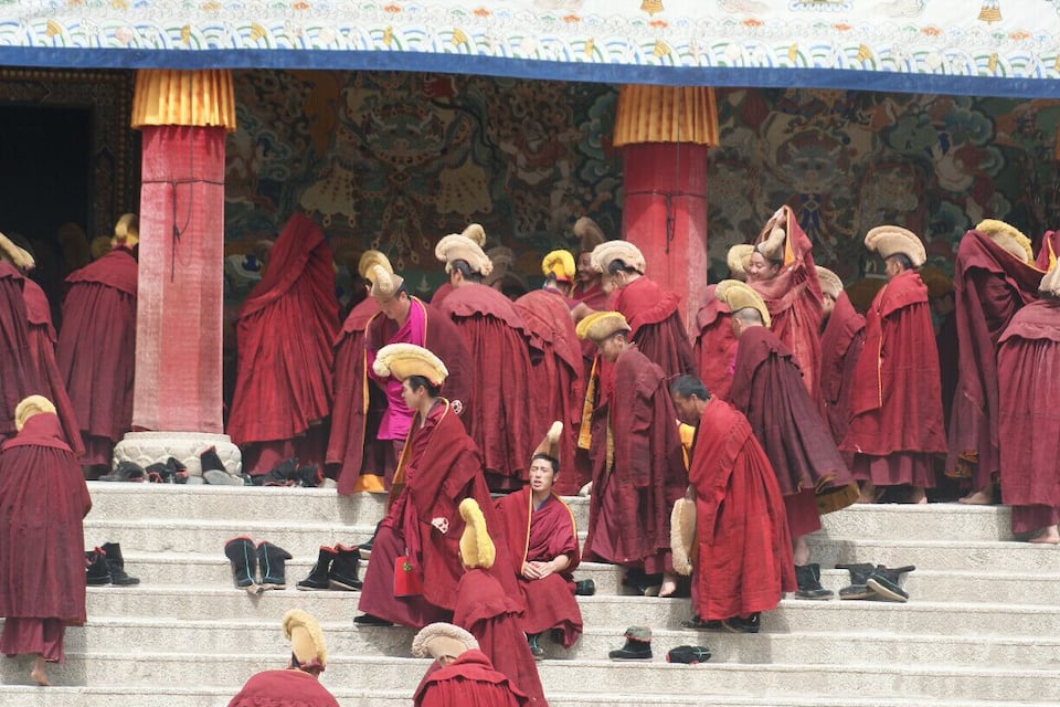 Monjes con túnicas rojas y sombreros amarillos en los escalones frente a un edificio con murales coloridos.