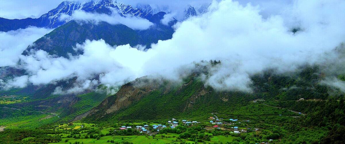 A mountainous landscape with a village nestled in the valley, surrounded by lush greenery and snow-capped peaks.