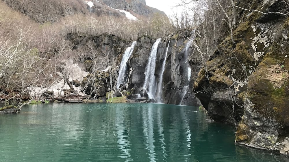 A waterfall cascading into a clear turquoise lake surrounded by rocky terrain and sparse vegetation.