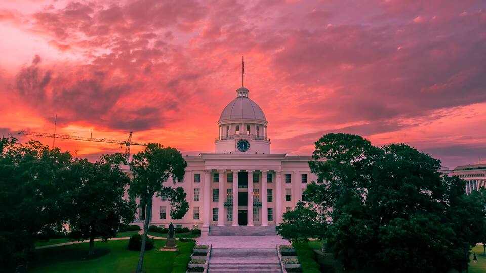 A large white building with a dome and columns, surrounded by trees and a sunset sky.