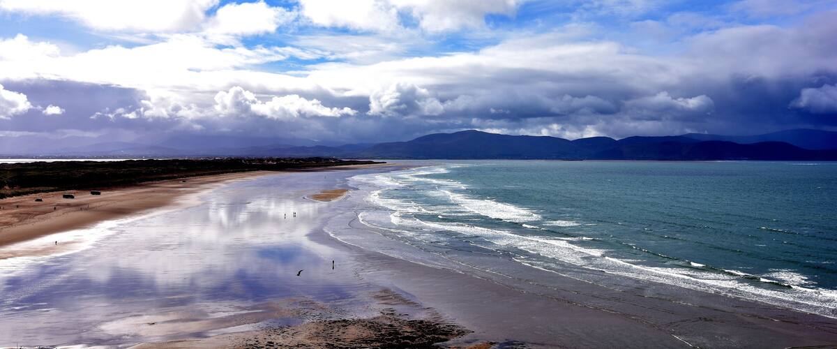 A beach with a calm sea, sandy shore, and distant mountains under a partly cloudy sky.