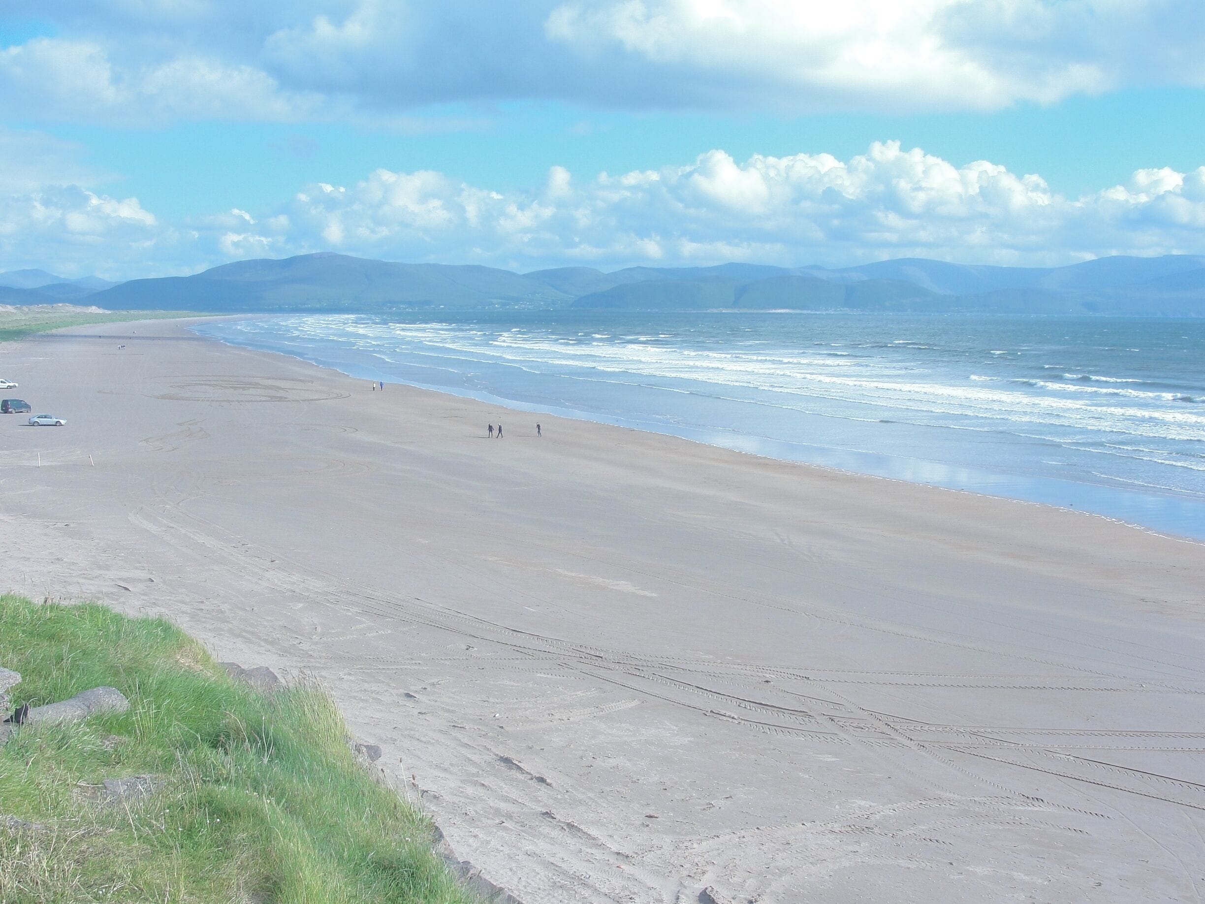 A wide sandy beach with gentle waves, a clear sky, and distant mountains.