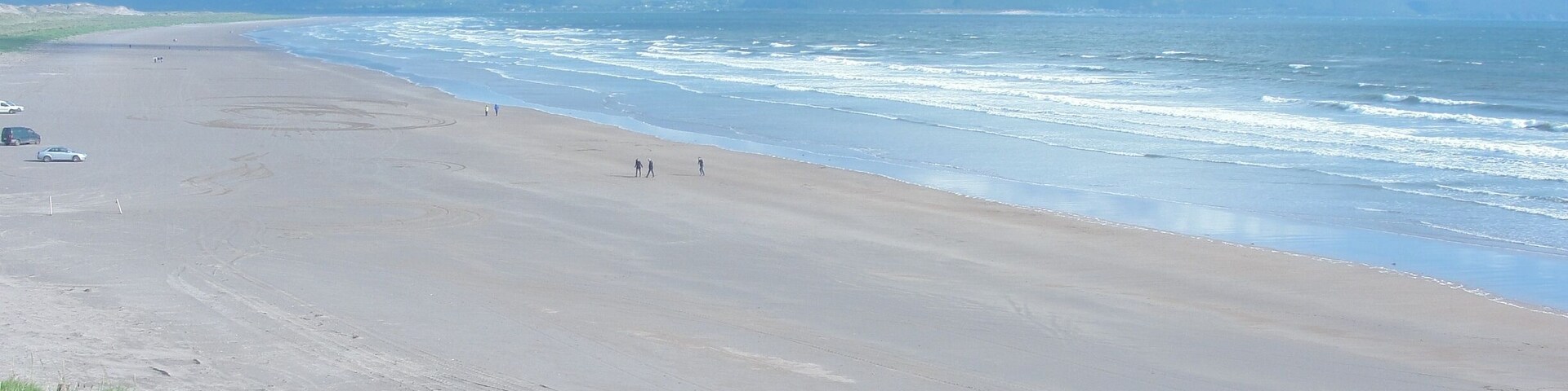 Una extensa playa de arena con olas suaves, cielo despejado y montañas a lo lejos.