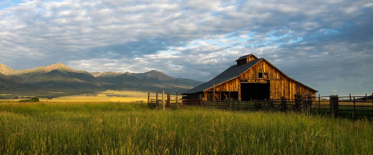 A rustic barn in a grassy field with mountains in the background.