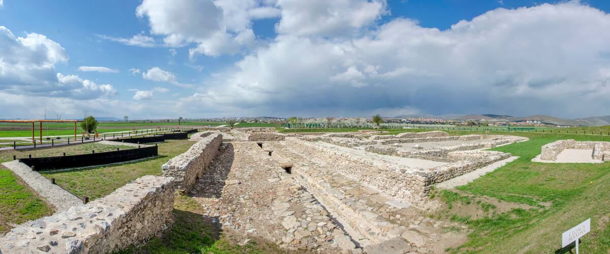 Ancient ruins with stone walls and pathways, surrounded by green fields and a clear sky.