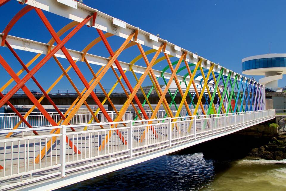 Un pont piétonnier aux couleurs vives, doté d’un motif géométrique, un bâtiment moderne en arrière-plan et une rivière en contrebas.