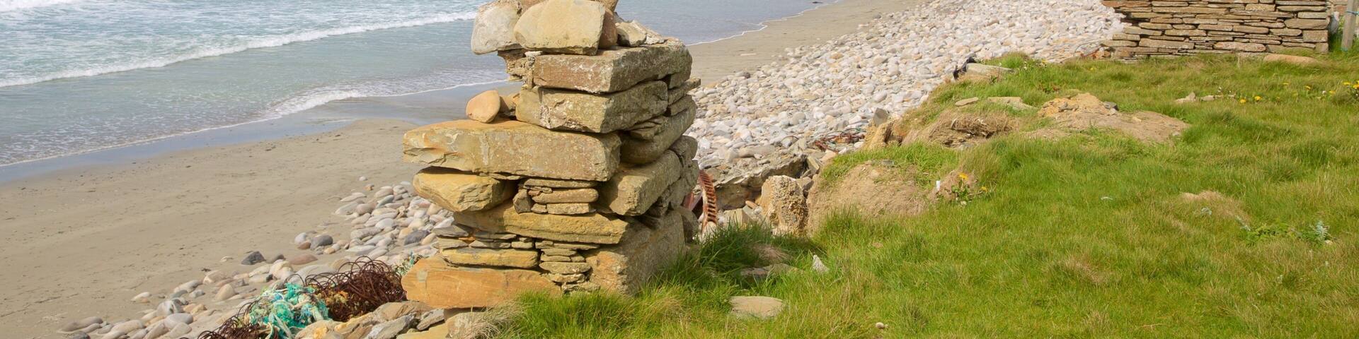 A coastal landscape with a stone wall, pebbly beach, and distant houses.