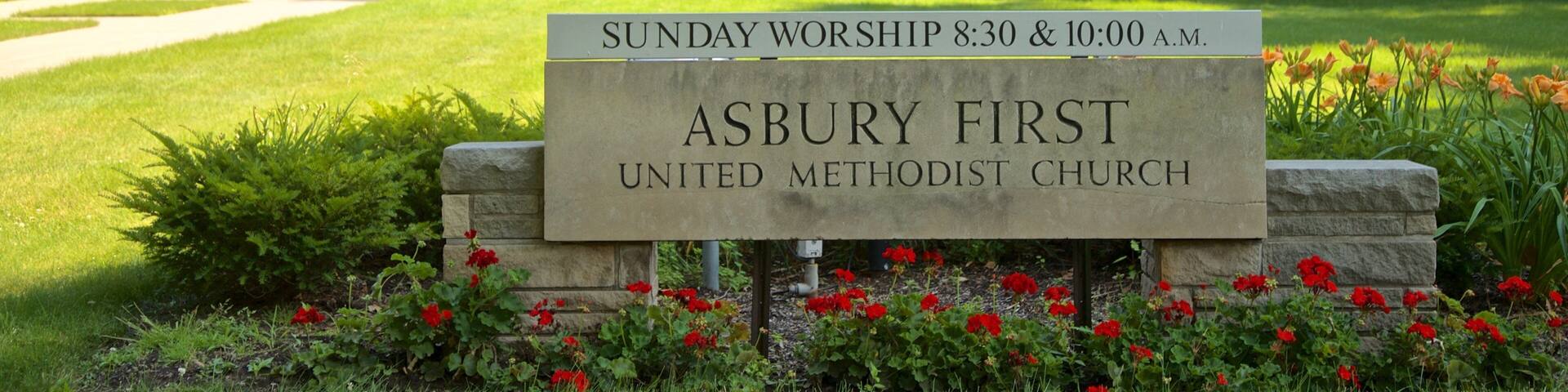 A stone sign in front of a church with a red door, surrounded by a well-maintained lawn and red flowers.
