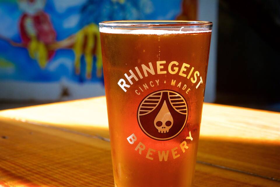 A glass of beer with a skull logo and text reading "RHINEGEIST CINCY MADE BREWERY" on a wooden surface.