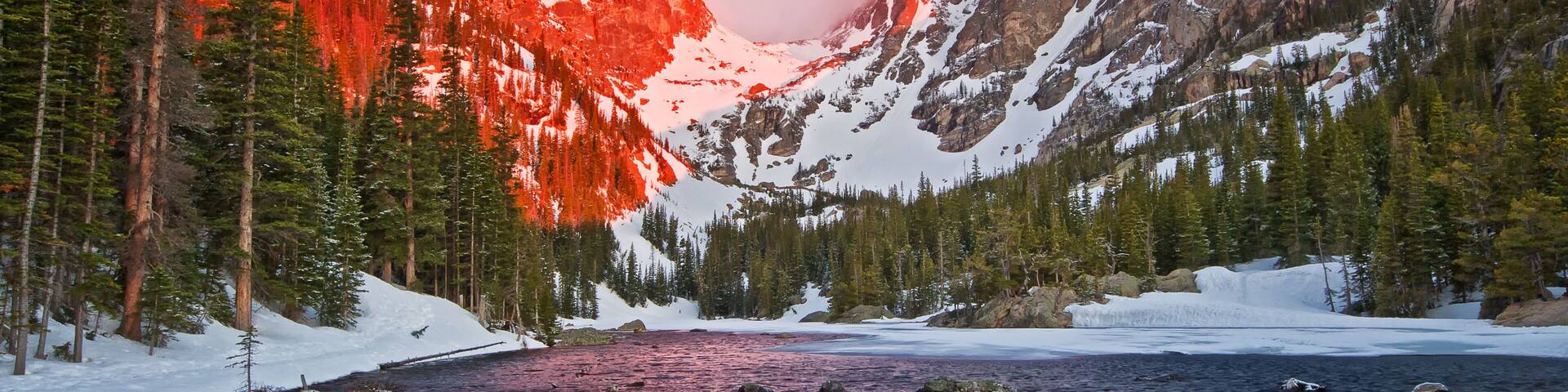 A mountain landscape with a river, snow-covered peaks, and dense forest.