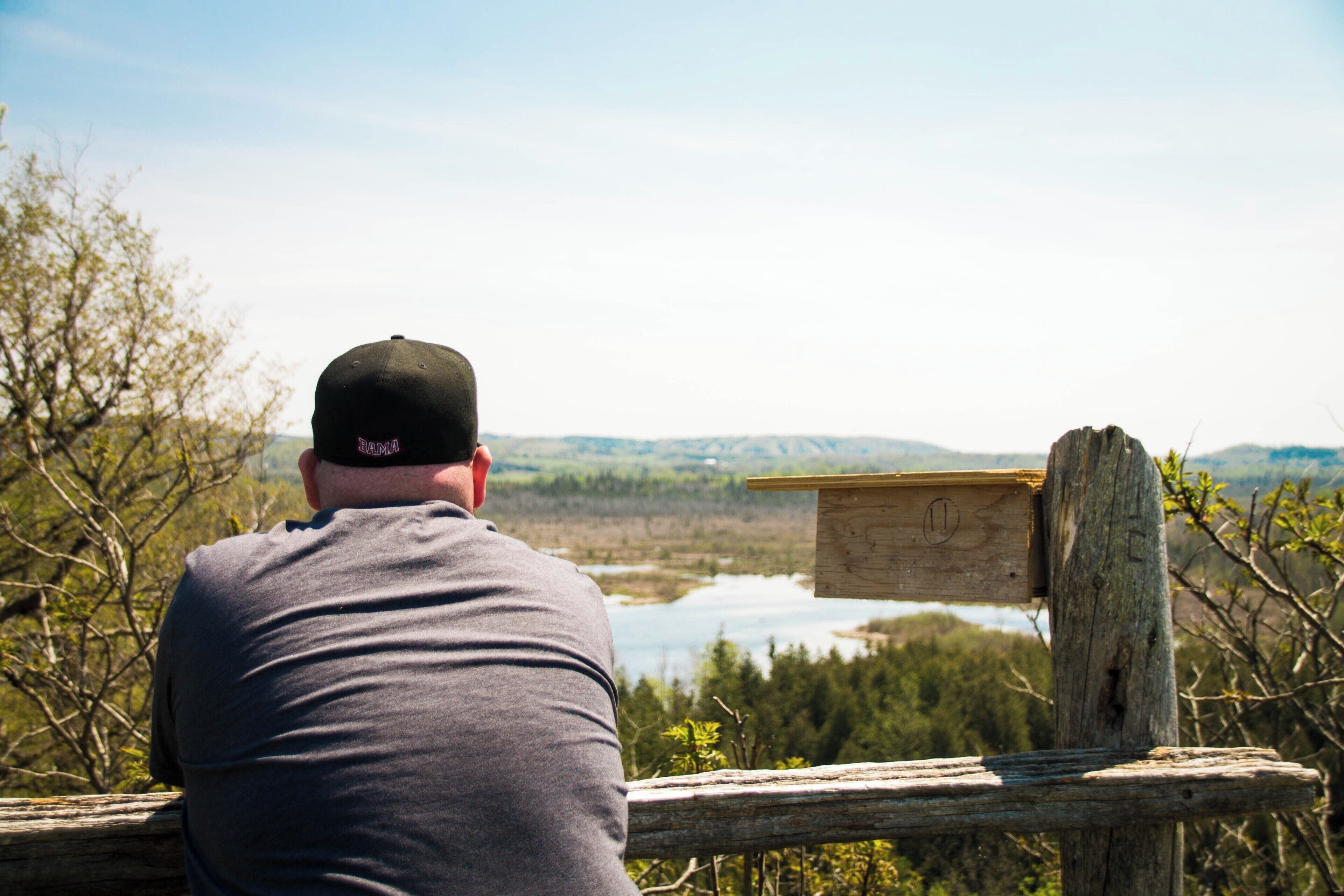 A man in a grey shirt and black cap overlooking a lake surrounded by trees and hills.