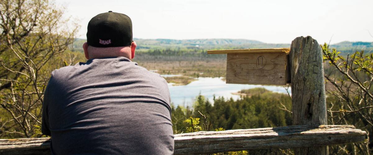 A man in a grey shirt and black cap overlooking a lake surrounded by trees and hills.