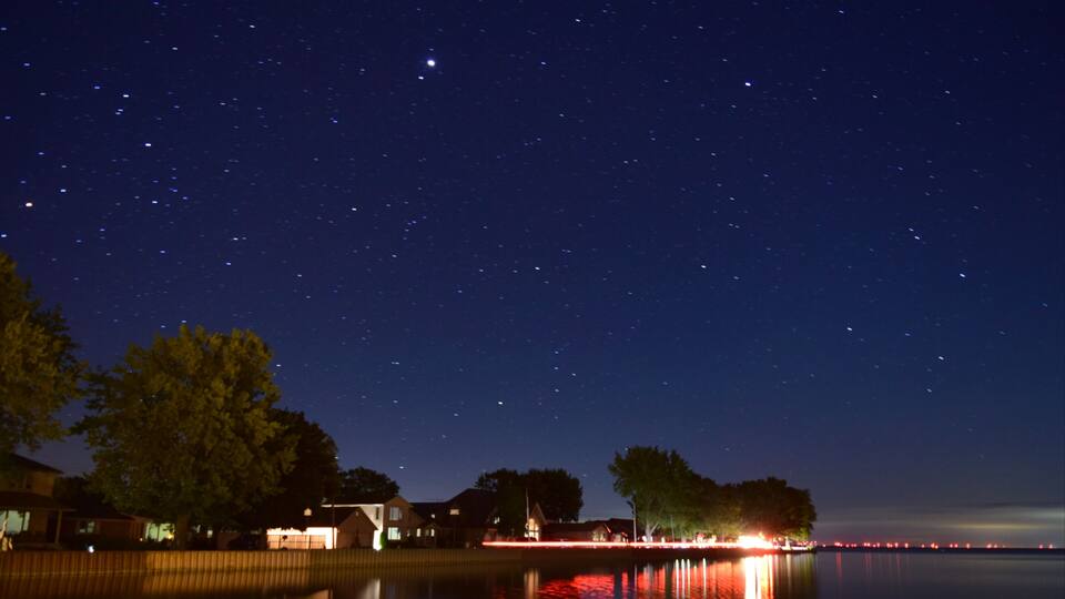 A night sky over a lakeside town with visible stars, houses, and a dock reflecting lights.
