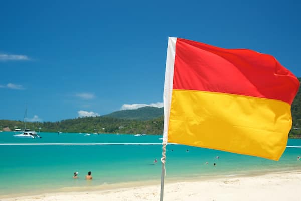 Uma praia com águas turquesa cristalinas, uma bandeira e barcos ao fundo.