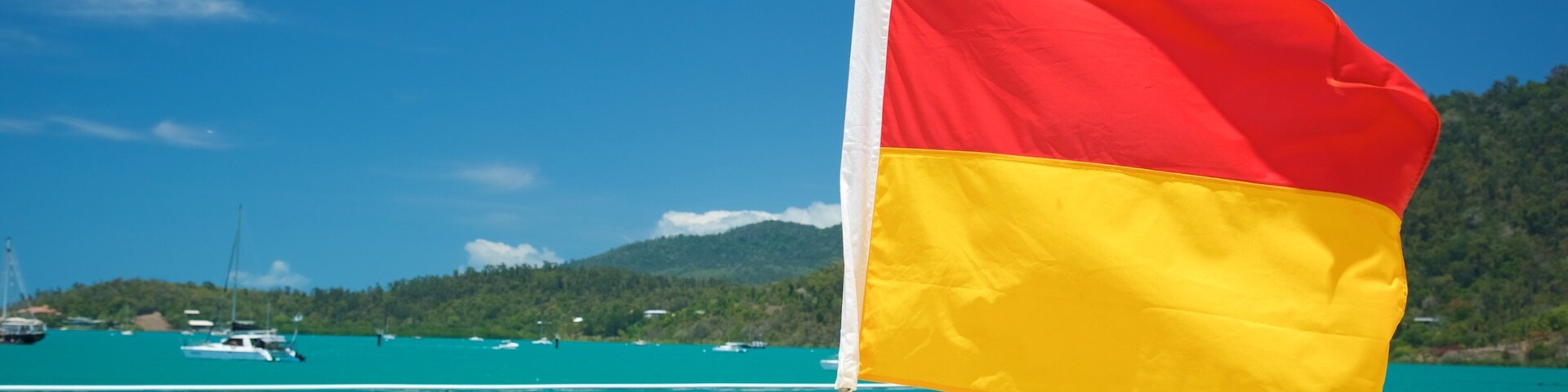 Una playa con agua turquesa cristalina, una bandera y barcos al fondo.