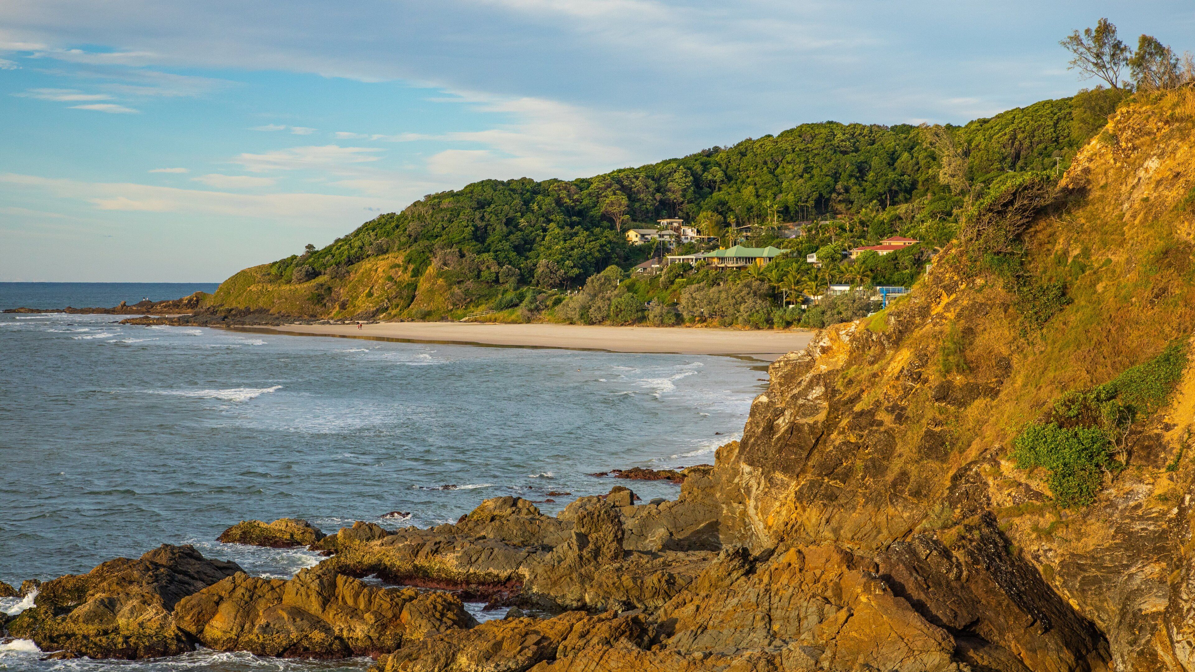 A coastal landscape with rocky cliffs, a sandy beach, and a hillside with houses.