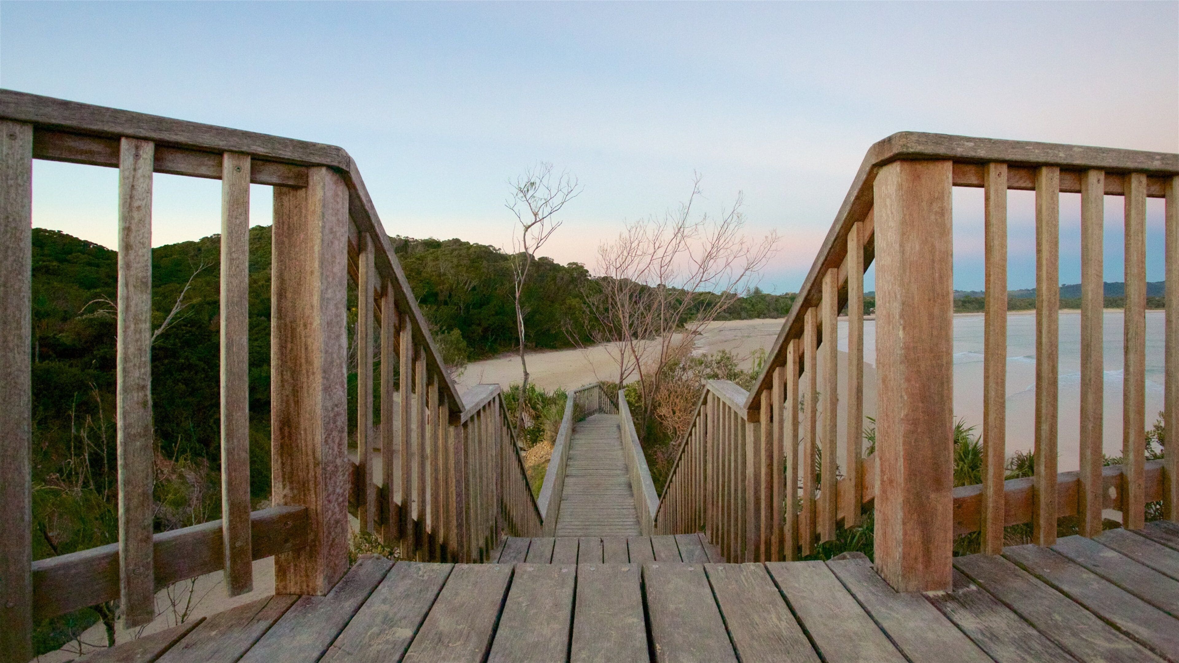 Un escalier en bois mène à une plage, d’où l’on peut admirer la vue sur l’eau et les collines en arrière-plan.