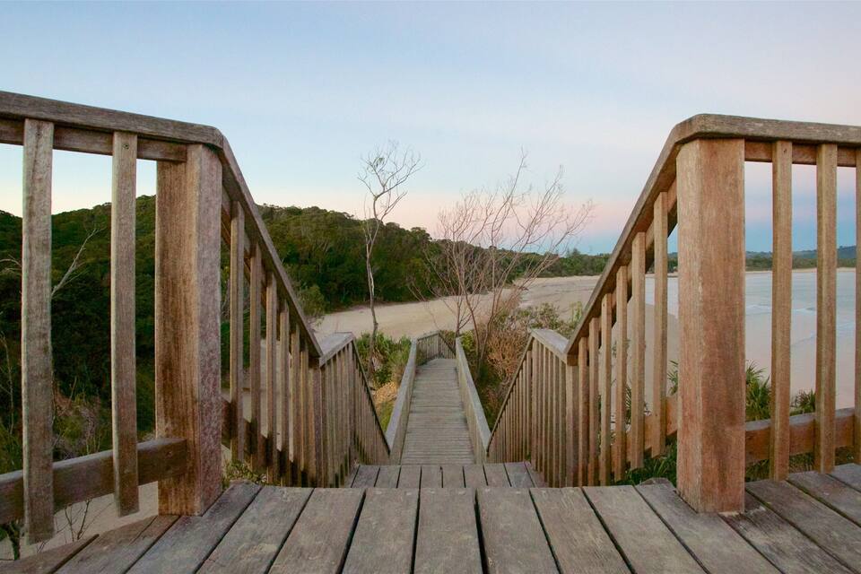 Una escalera de madera que conduce a una playa, con vista al agua y colinas al fondo.