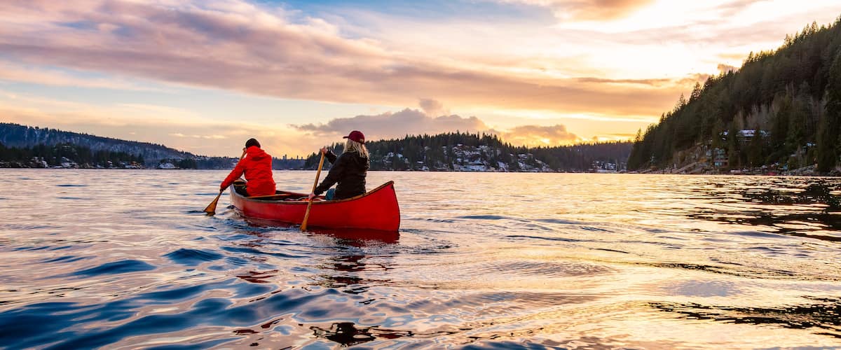 A red canoe on a calm lake with a forested shoreline and a sunset in the background.