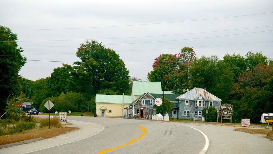 A rural road with a yellow curve sign, a blue building with a green roof, and a sign welcoming visitors to Phillips.