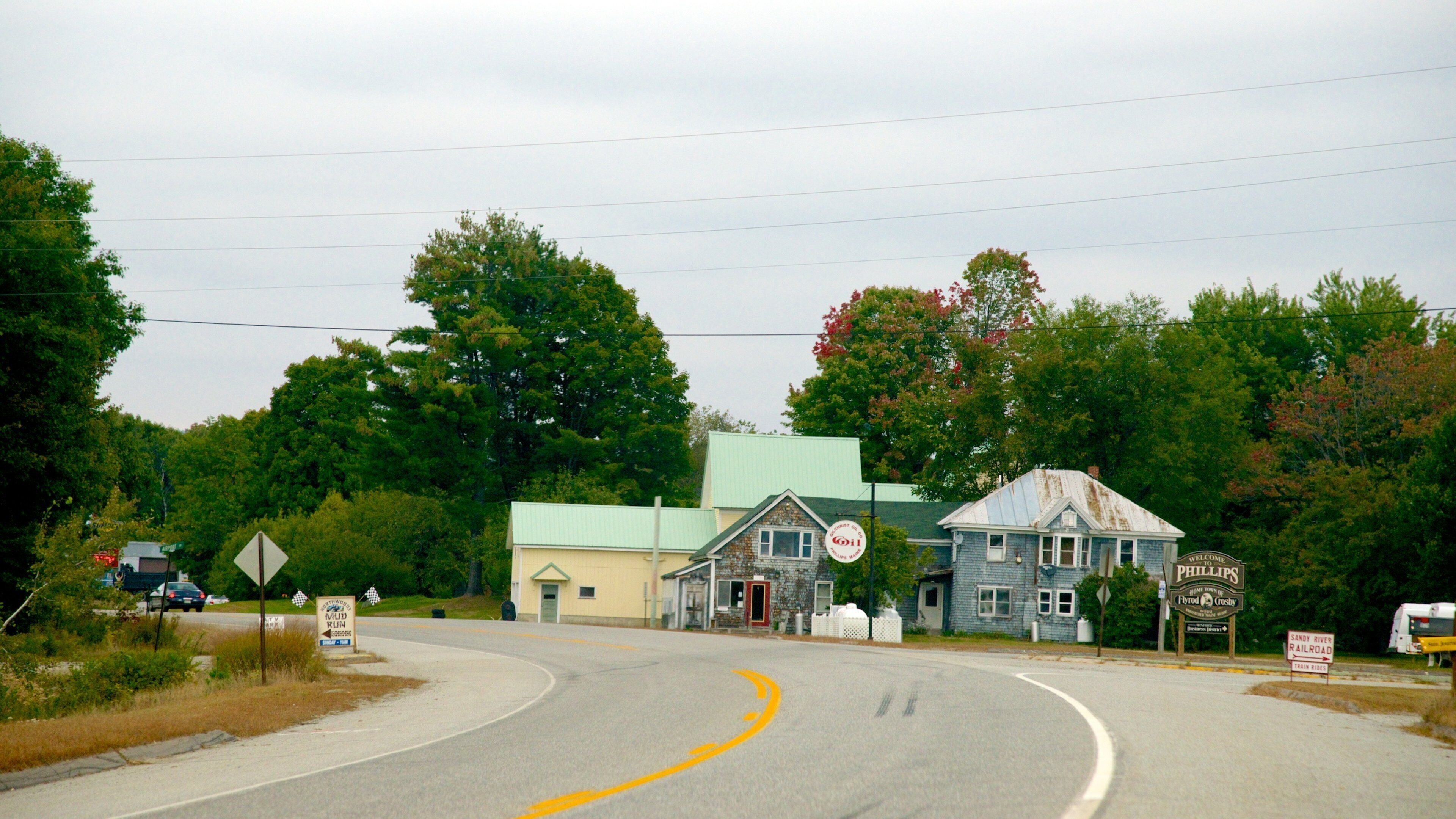 A rural road with a yellow curve sign, a blue building with a green roof, and a sign welcoming visitors to Phillips.