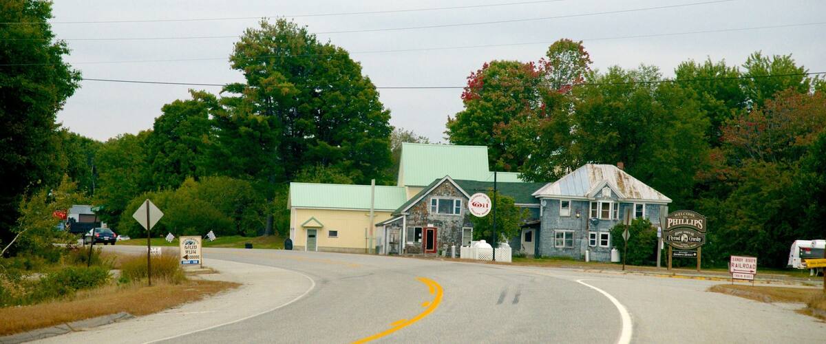 A rural road with a yellow curve sign, a blue building with a green roof, and a sign welcoming visitors to Phillips.